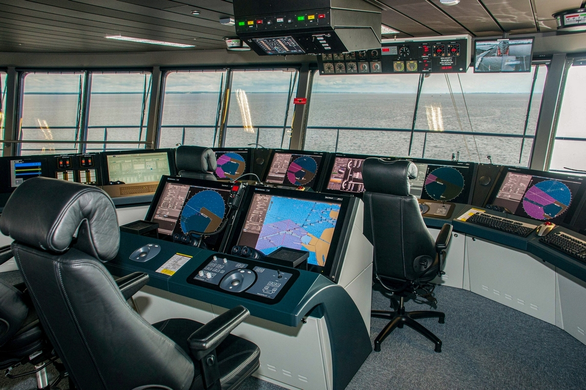 Ship bridge control room with ECDIS displays and navigation consoles showing charts and radar, viewed from behind the operator chairs.