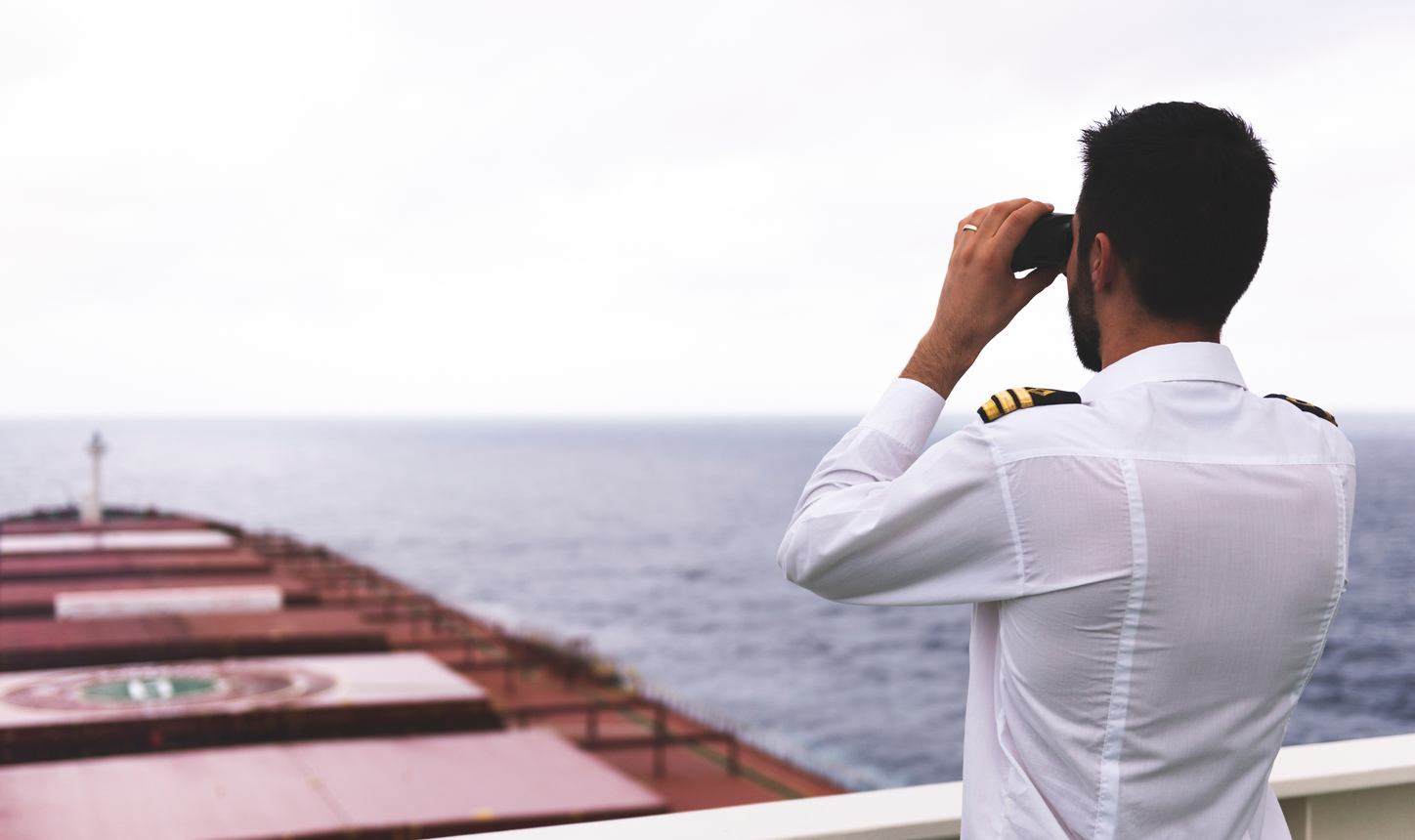 Ship officer on a cargo vessel’s deck looking out to sea through binoculars.