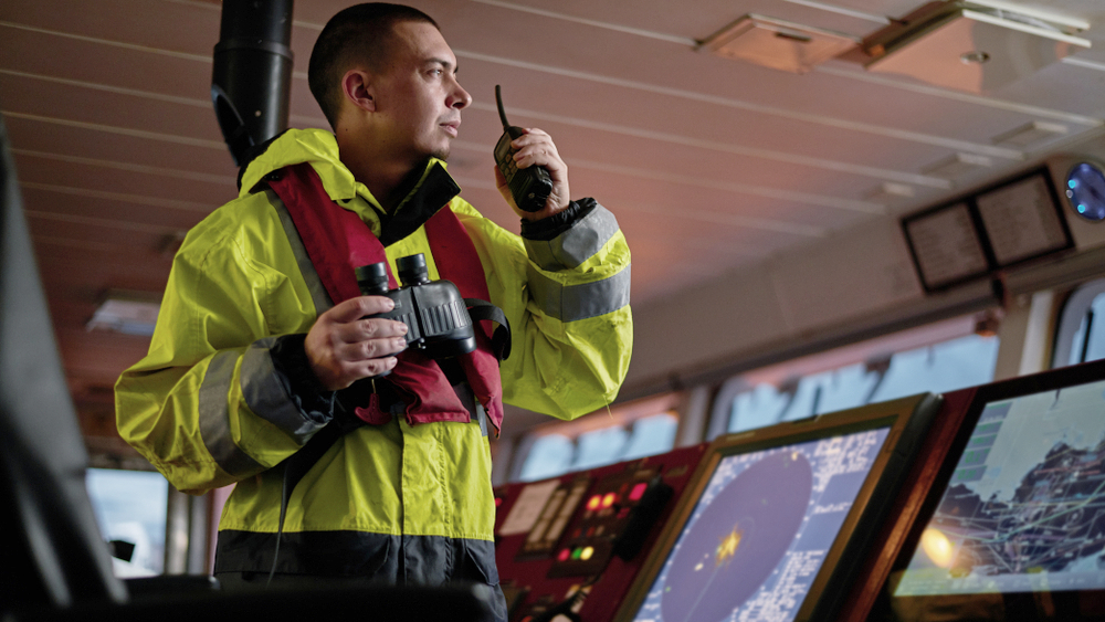 rew member on a ship’s bridge wearing a high-visibility jacket and life vest, holding binoculars and speaking into a handheld radio beside navigation displays.
